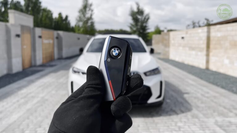 A close-up of a BMW key fob held by a gloved hand, with a white BMW SUV in the background