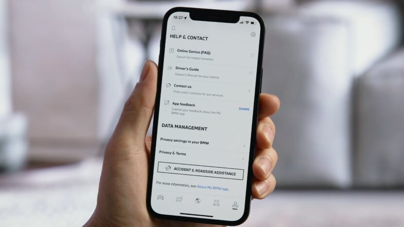 Hand holding a smartphone showing the My BMW app's Help & Contact screen with support options including FAQs, Driver’s Guide, contact details, app feedback, privacy settings, and roadside assistance