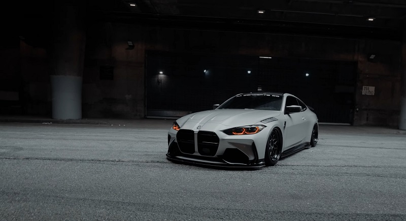 A matte grey BMW M4 Coupe (G82) with black wheels parked in a dimly lit urban garage