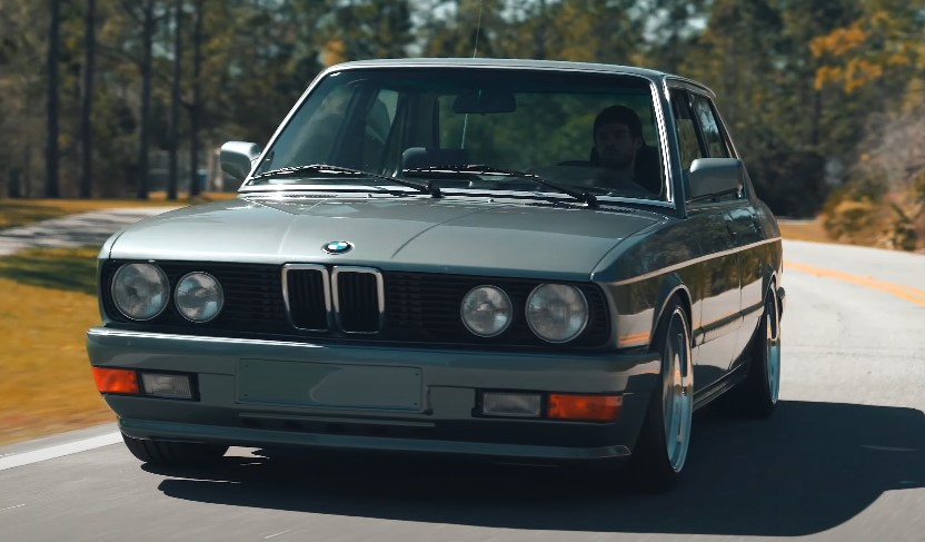A sleek gray E28 BMW coupe glides along a sun-drenched road