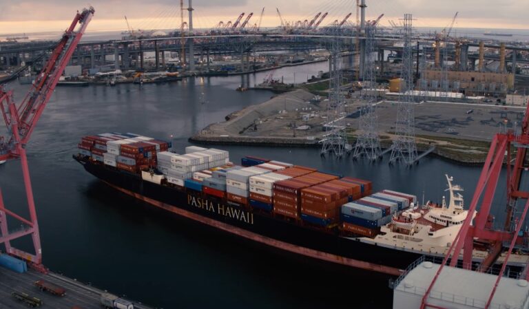 A large container ship, the Pasha Hawaii, navigates a busy port, laden with cargo