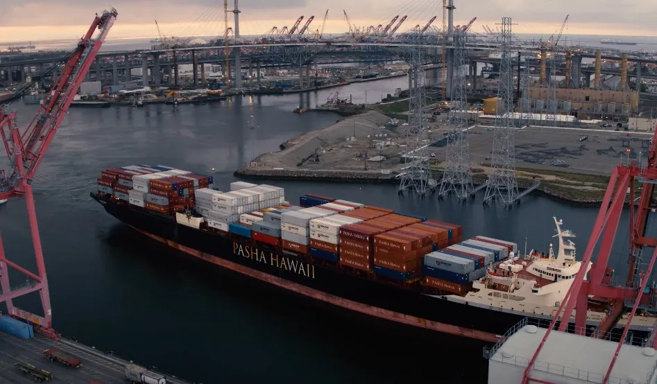 A large container ship, the Pasha Hawaii, navigates a busy port, laden with cargo