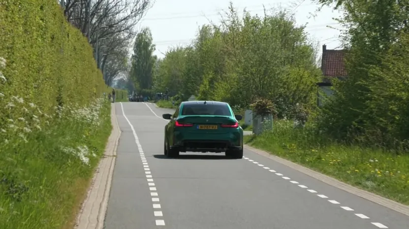 A green BMW M3 drives down a quiet country road lined with trees and grass