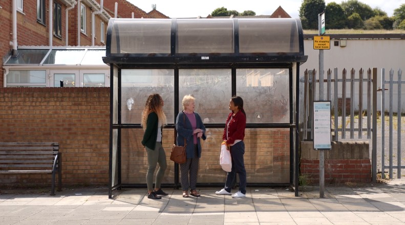 Three women wait for the bus, engaging in conversation under the bus shelter