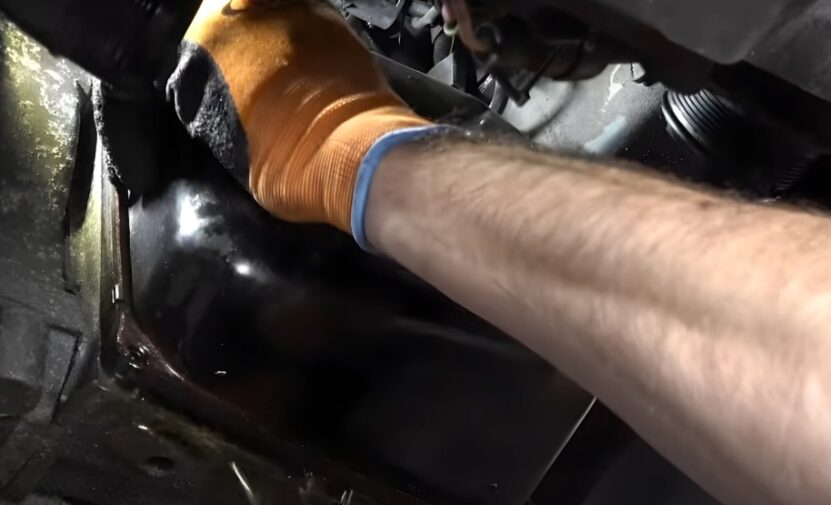 A hand in protective orange work gloves manipulates a dark component beneath the vehicle