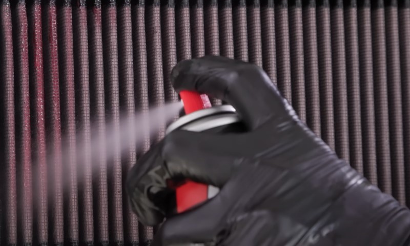 Black-gloved hand applying aerosol spray to a pleated, reddish-brown air filter element