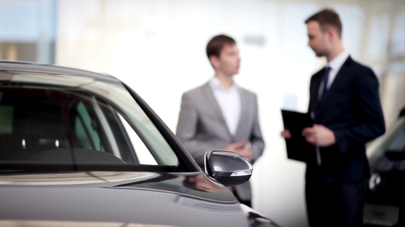 Two men in suits talking near a car inside a dealership showroom