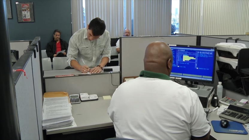 Person filling out paperwork at a DMV counter while others wait in the background