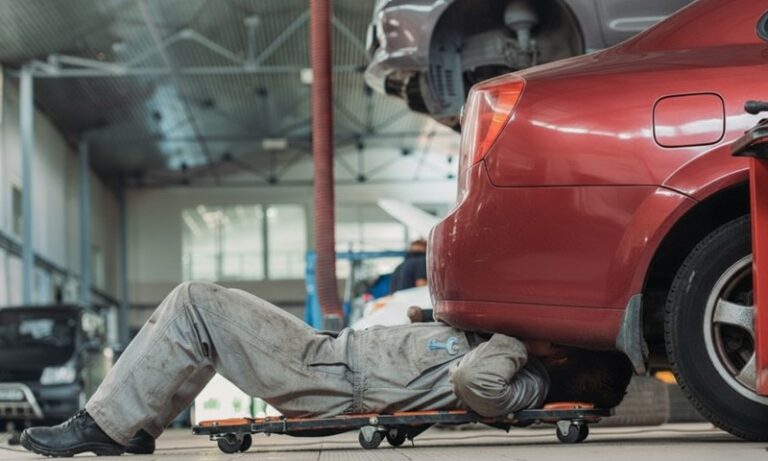 A mechanic in overalls lies on a creeper beneath a red car in a garage