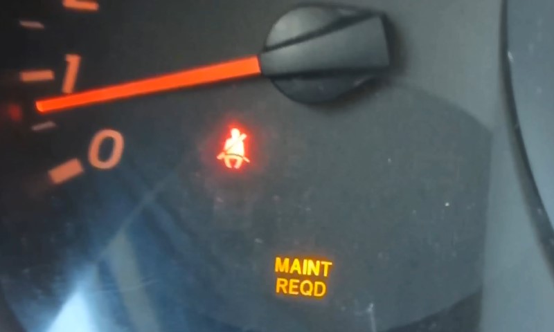 Close-up of a car dashboard showing a red seatbelt warning light and an orange "Maint Reqd" indicator