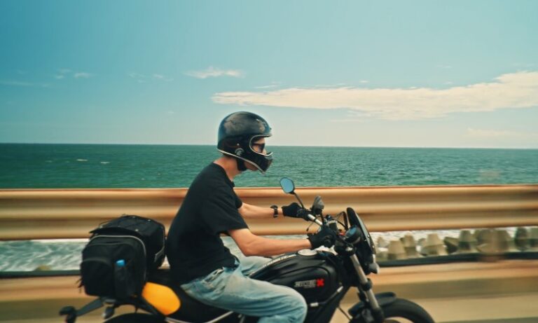 A person rides a motorcycle along a coastal road with the ocean and blue sky in the background