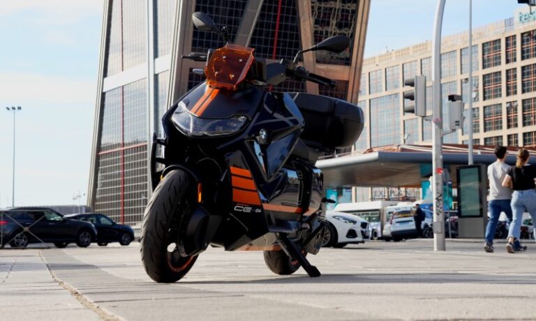 A sleek black and orange electric scooter is parked on a city sidewalk, with modern glass buildings in the background